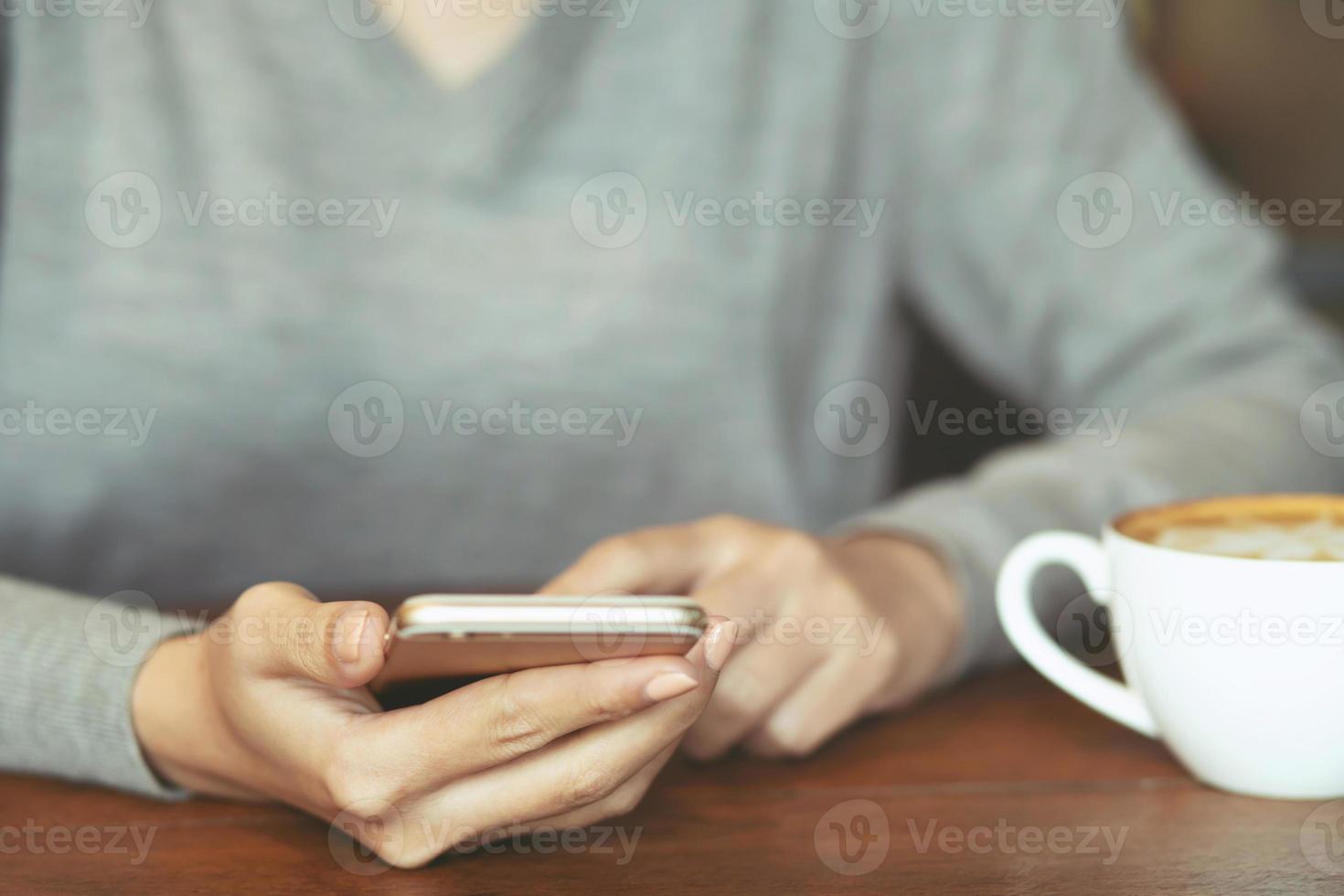 Feche as mãos da mulher segurando usando a tela de espaço de cópia em branco do telefone móvel inteligente vazia para sua mensagem de texto de publicidade na vista superior e bebendo uma xícara de café no fundo da mesa de madeira no café. foco suave foto