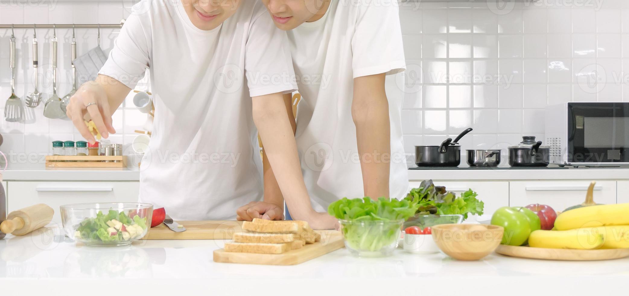 meio retrato asiático jovem lgbt masculino casal feliz cozinhar salada juntos no fundo branco da cozinha em casa. conceito de vida doméstica. foco seletivo. foto