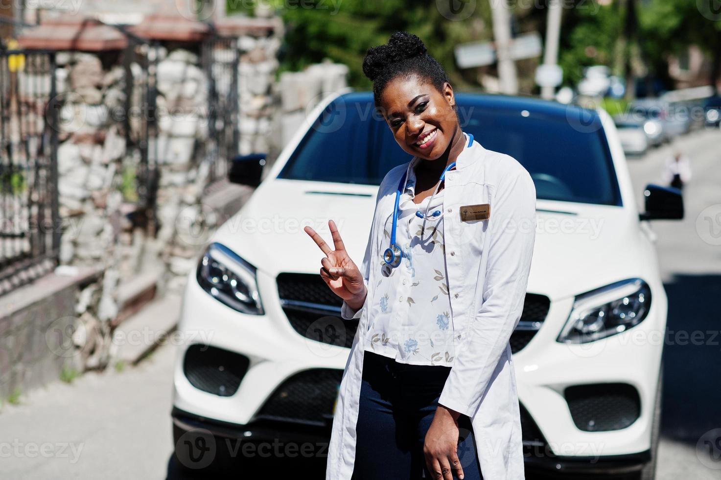 médico americano africano fêmea no jaleco com estetoscópio posou ao ar livre contra carro suv branco. foto