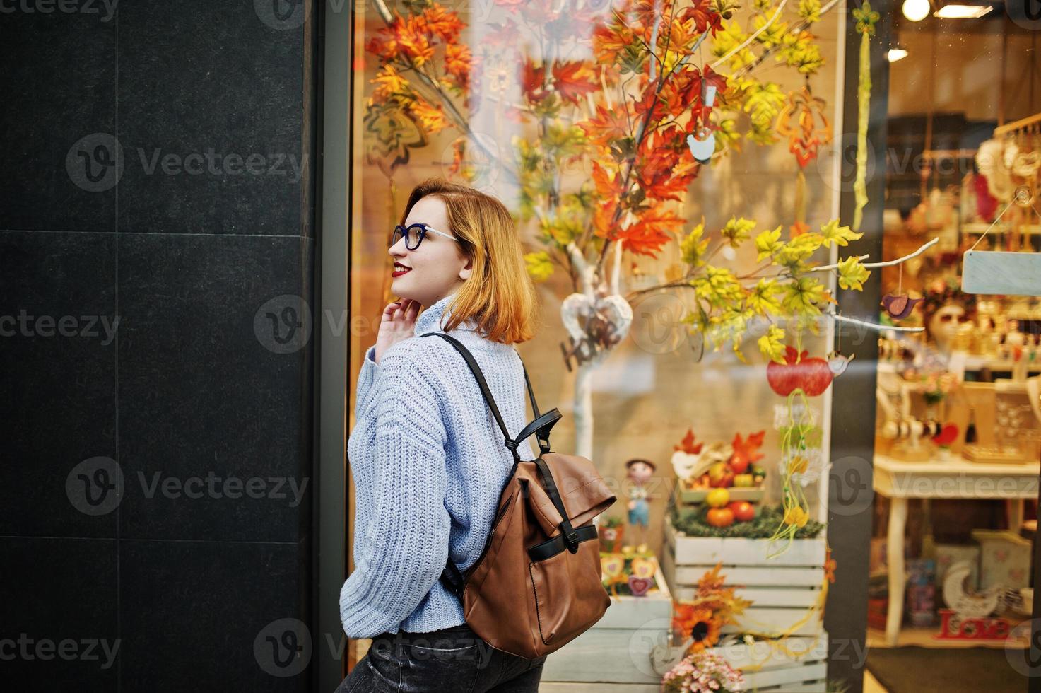 alegre jovem ruiva bonita de óculos, suéter de lã azul quente com mochila posou ao ar livre contra folhas de outono decorações de árvores na loja. foto