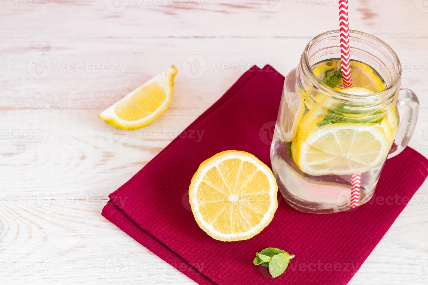 frasco de vidro de limonada caseira com limões, hortelã e palha de papel vermelho em guardanapo de pano e fundo de madeira. bebida refrescante de verão. foto