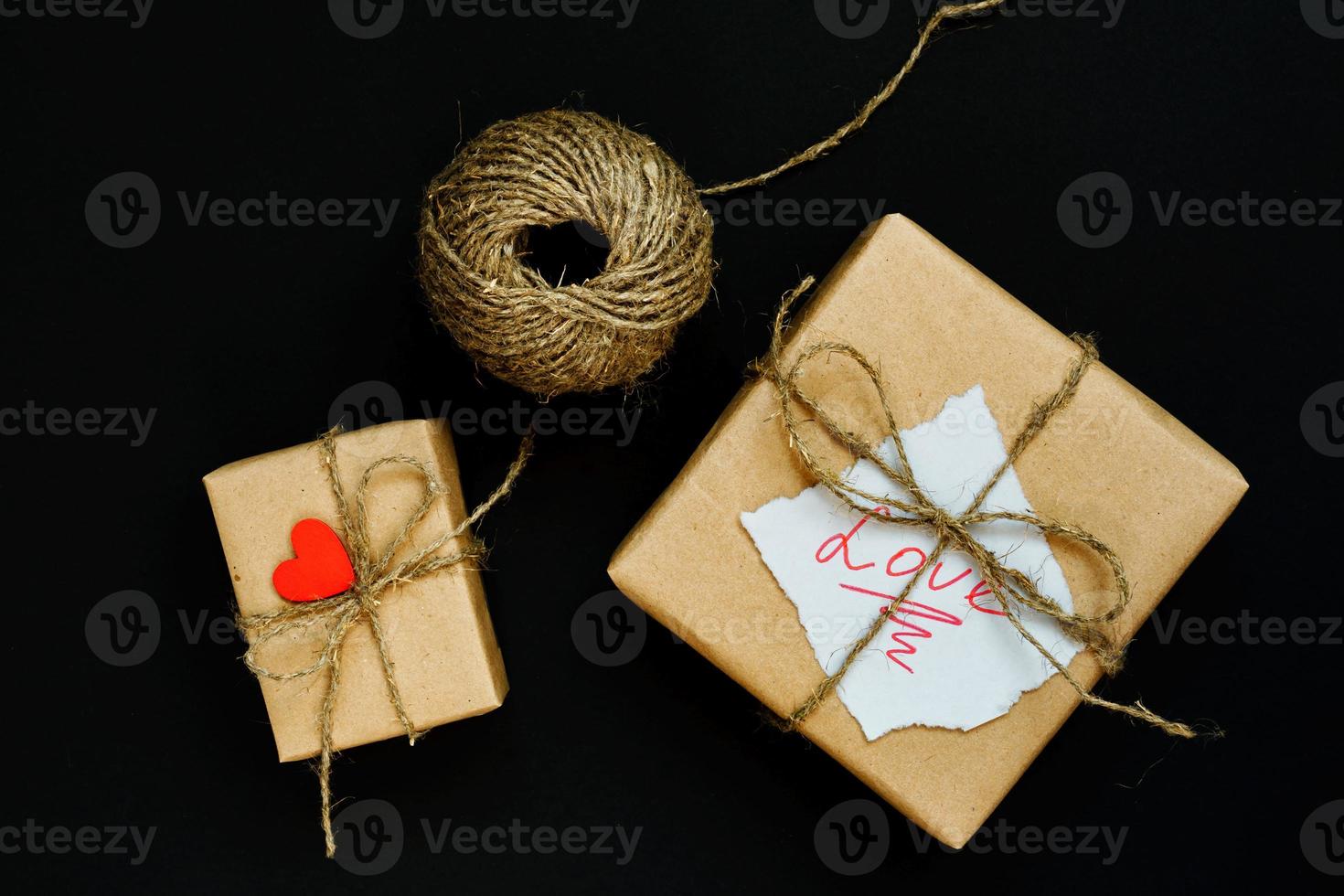 caixa de presente artesanal embrulhada em papel ofício com coração de madeira vermelho, corda e laço em fundo preto. vista superior, configuração plana foto