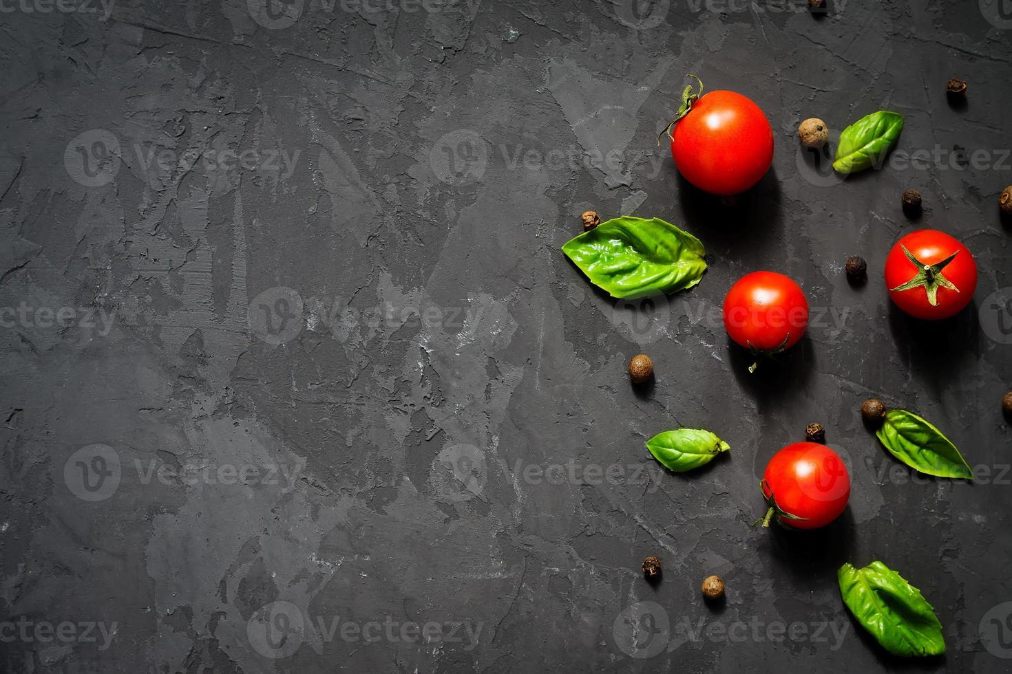 suculentos tomates cereja maduros com folhas de manjericão verde e pimenta preta sobre fundo preto. conceito de comida saudável. vista superior, copie o espaço foto