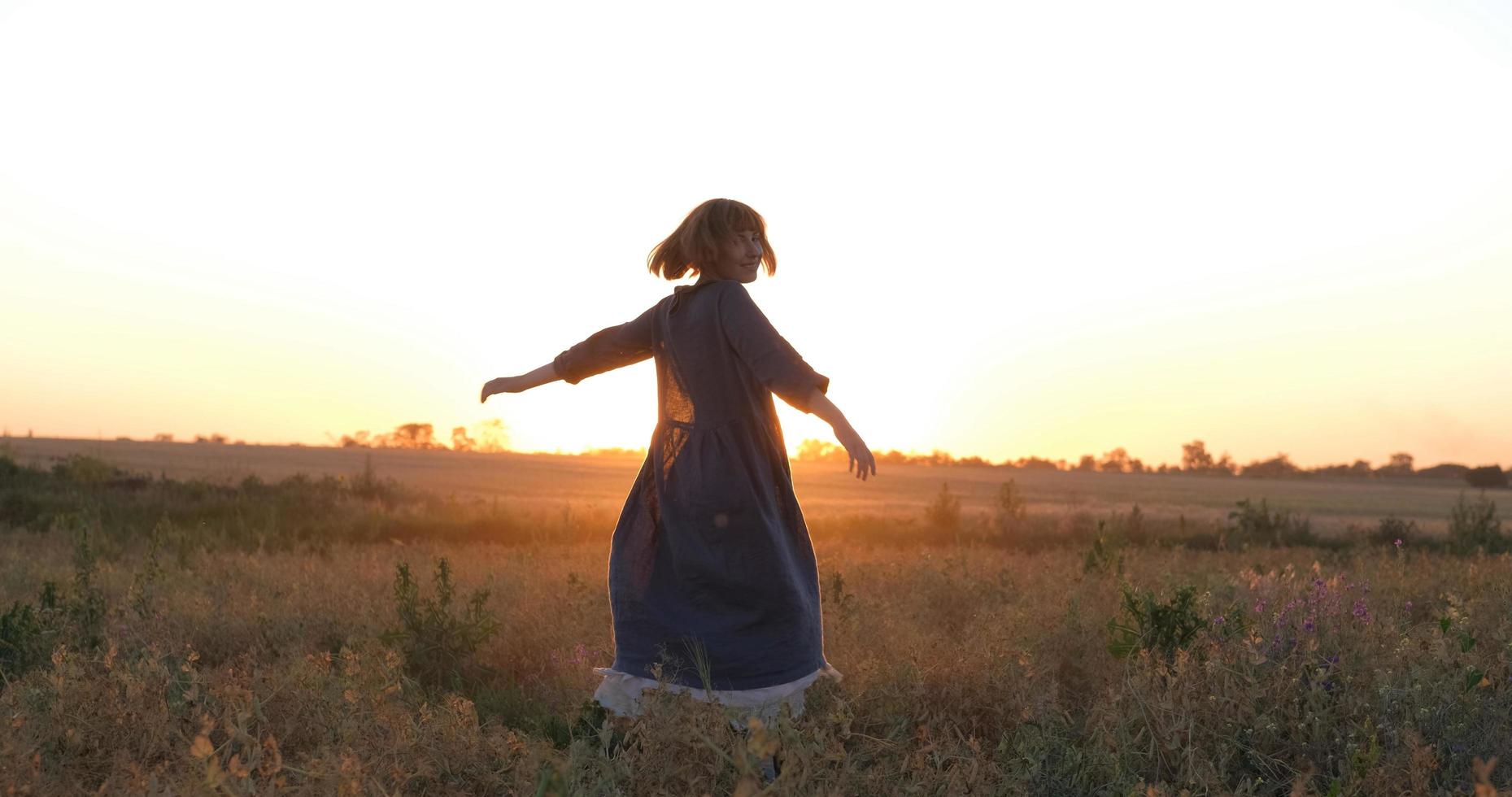 jovem ruiva em lindo vestido boho relaxando no campo durante o pôr do sol nevoento, feminino ao ar livre com buquê nas mãos foto