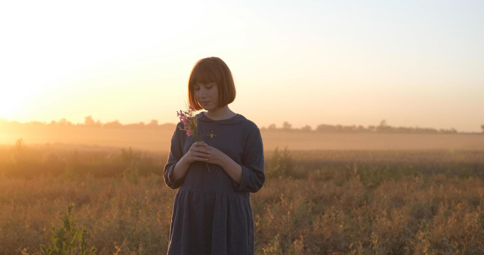 jovem ruiva em lindo vestido boho relaxando no campo durante o pôr do sol nevoento, feminino ao ar livre com buquê nas mãos foto