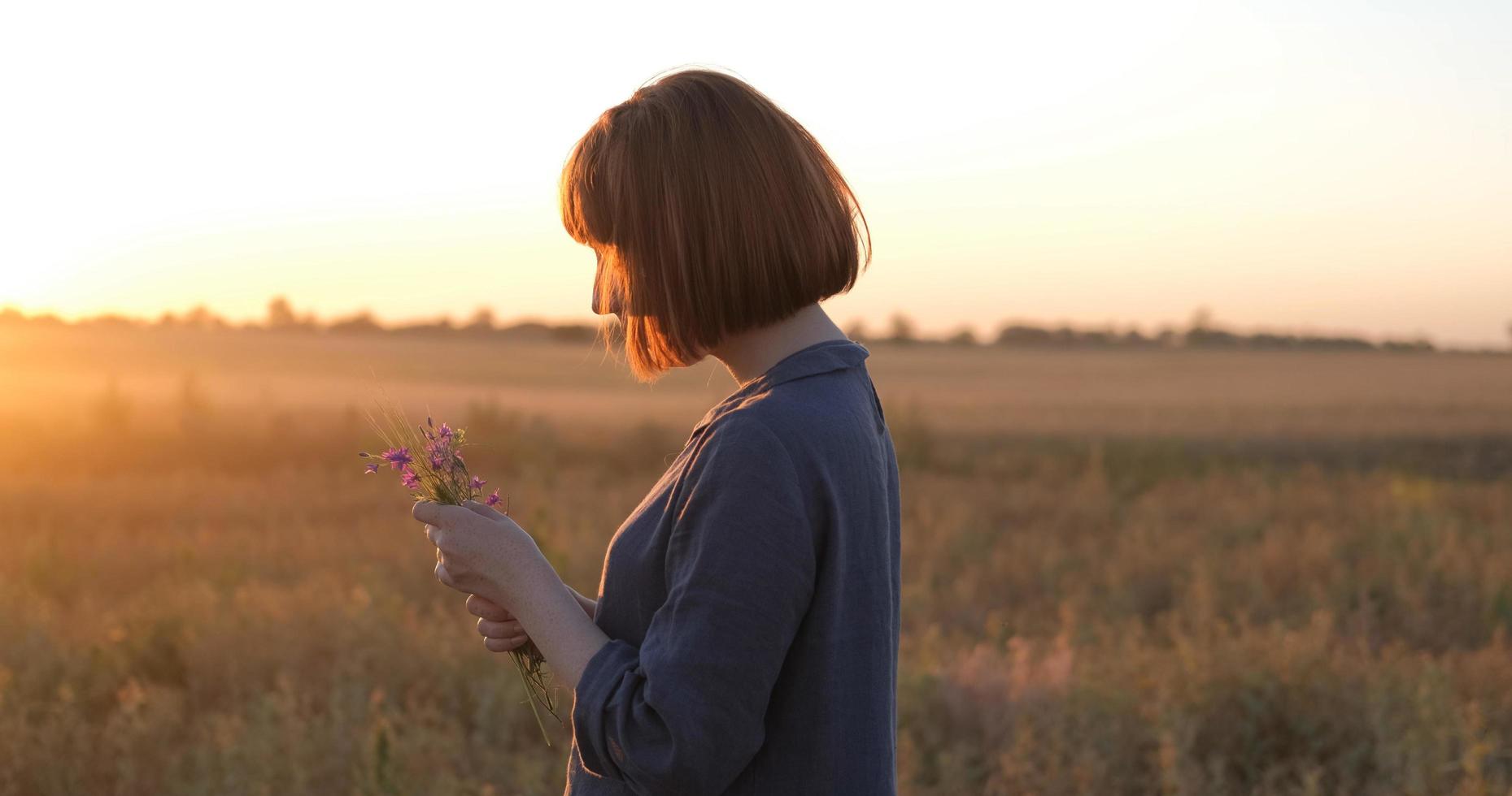 jovem ruiva em lindo vestido boho relaxando no campo durante o pôr do sol nevoento, feminino ao ar livre com buquê nas mãos foto