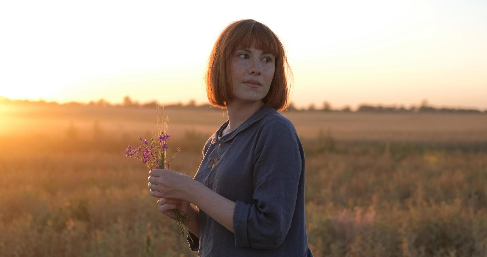 jovem ruiva em lindo vestido boho relaxando no campo durante o pôr do sol nevoento, feminino ao ar livre com buquê nas mãos foto