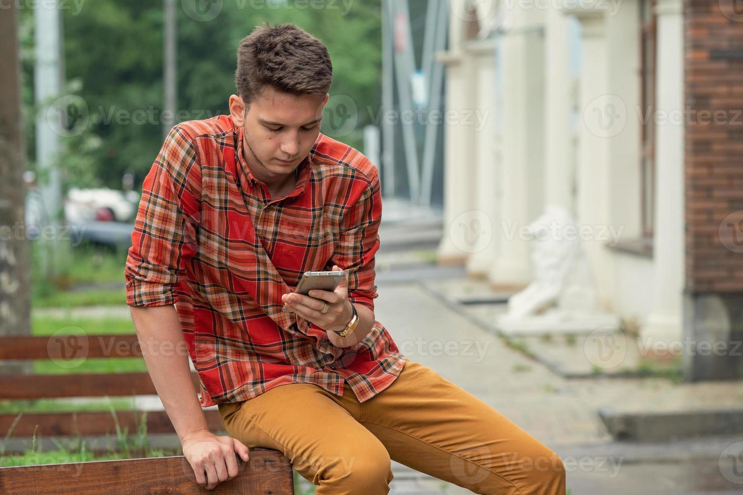 um jovem de camisa xadrez senta-se em uma grade de madeira, segura um telefone nas mãos e digita uma mensagem foto