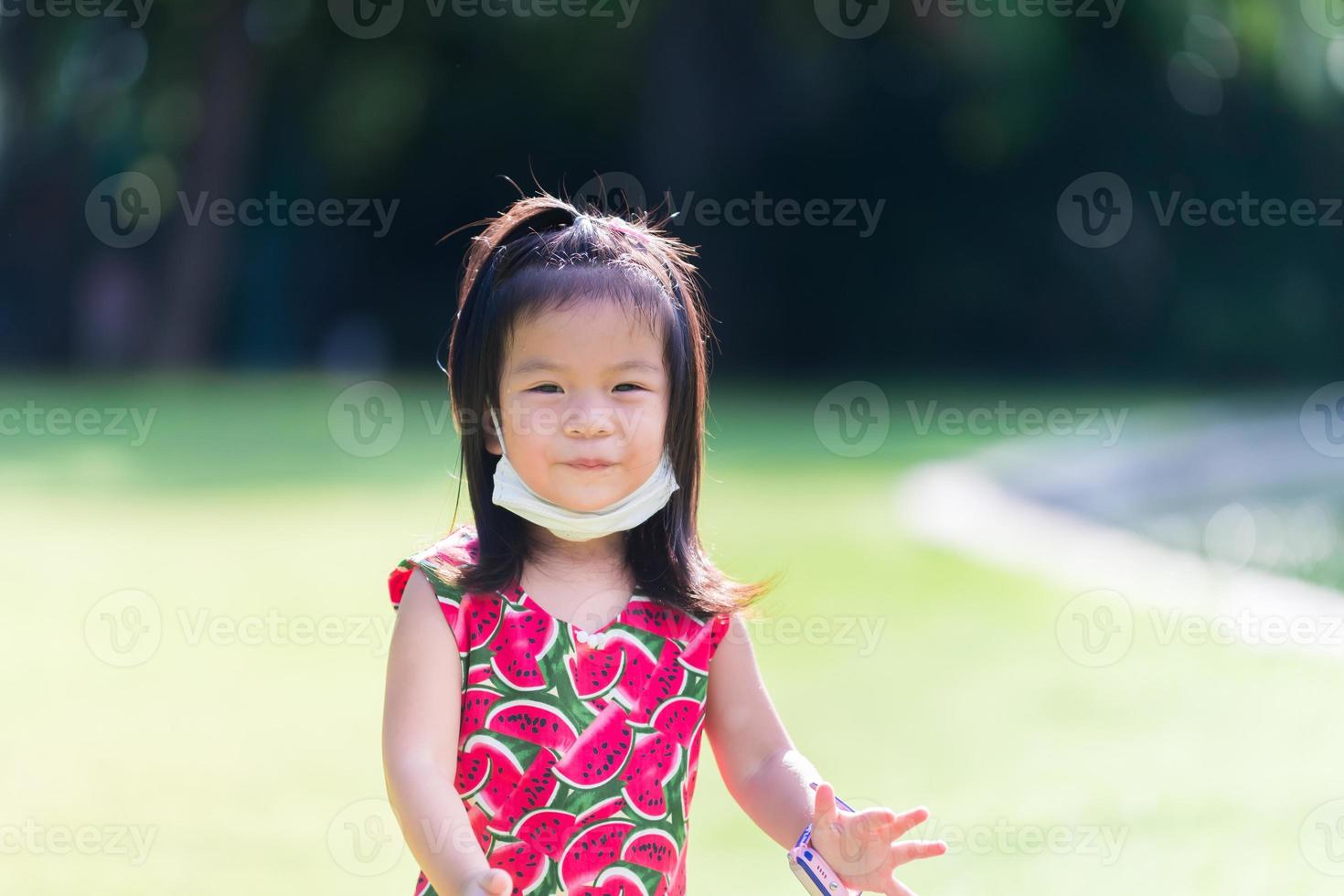 sorriso doce linda garota asiática. criança leva máscara facial sob o queixo. fundo verde brilhante do gramado. verão ou primavera. criança feliz vestida de vermelho olhou para a câmera, no sol quente. crianças de 3 a 4 anos. foto