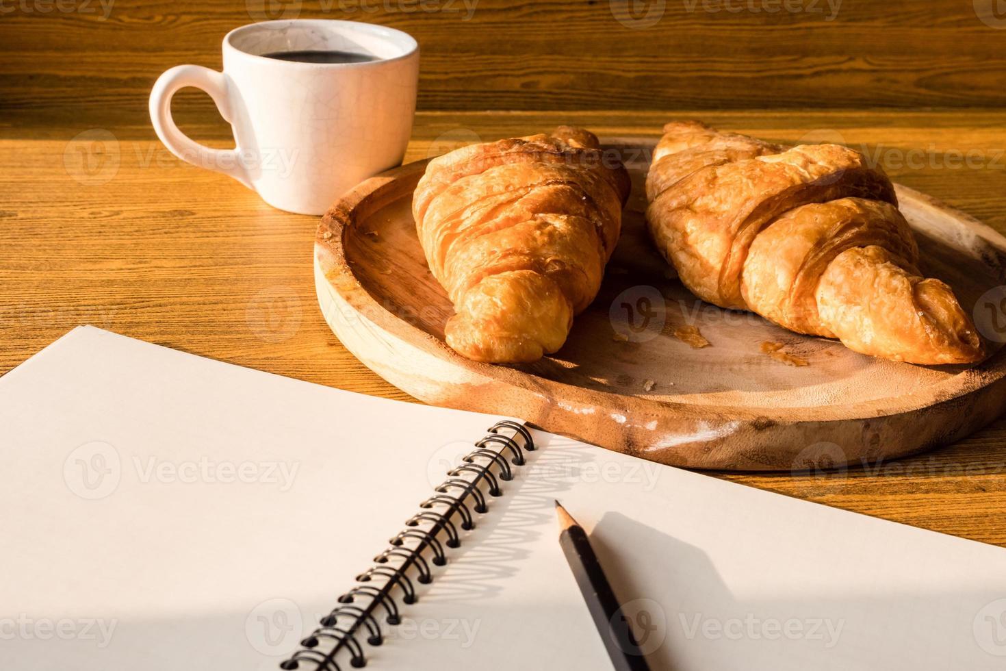 croissant com xícara de café, caderno e lápis no fundo da mesa de madeira. foto