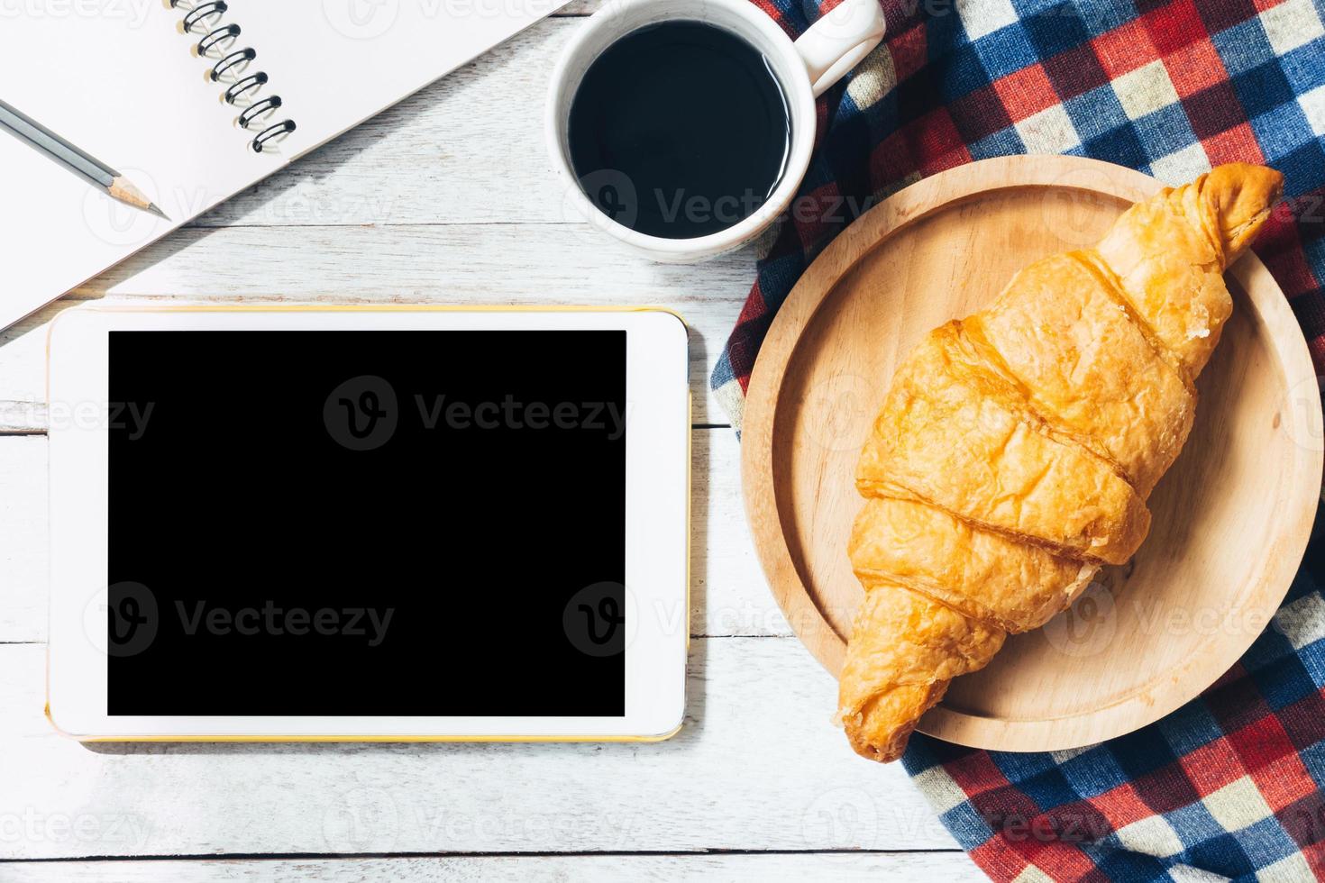 vista superior do tablet com croissant e café no fundo da mesa de madeira. foto