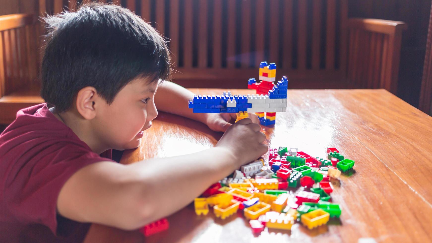 menino asiático está ficando criativo com a montagem de tijolos de plástico coloridos em robôs e aviões em uma mesa de madeira feliz e divertido no conceito de criadores home.kid. foto