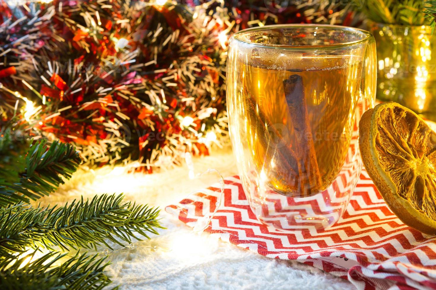 copo de vidro transparente de parede dupla com chá quente e paus de canela em cima da mesa com decoração de natal. atmosfera de ano novo, fatia de laranja seca, guirlanda e enfeites, ramo de abeto, aconchegante foto