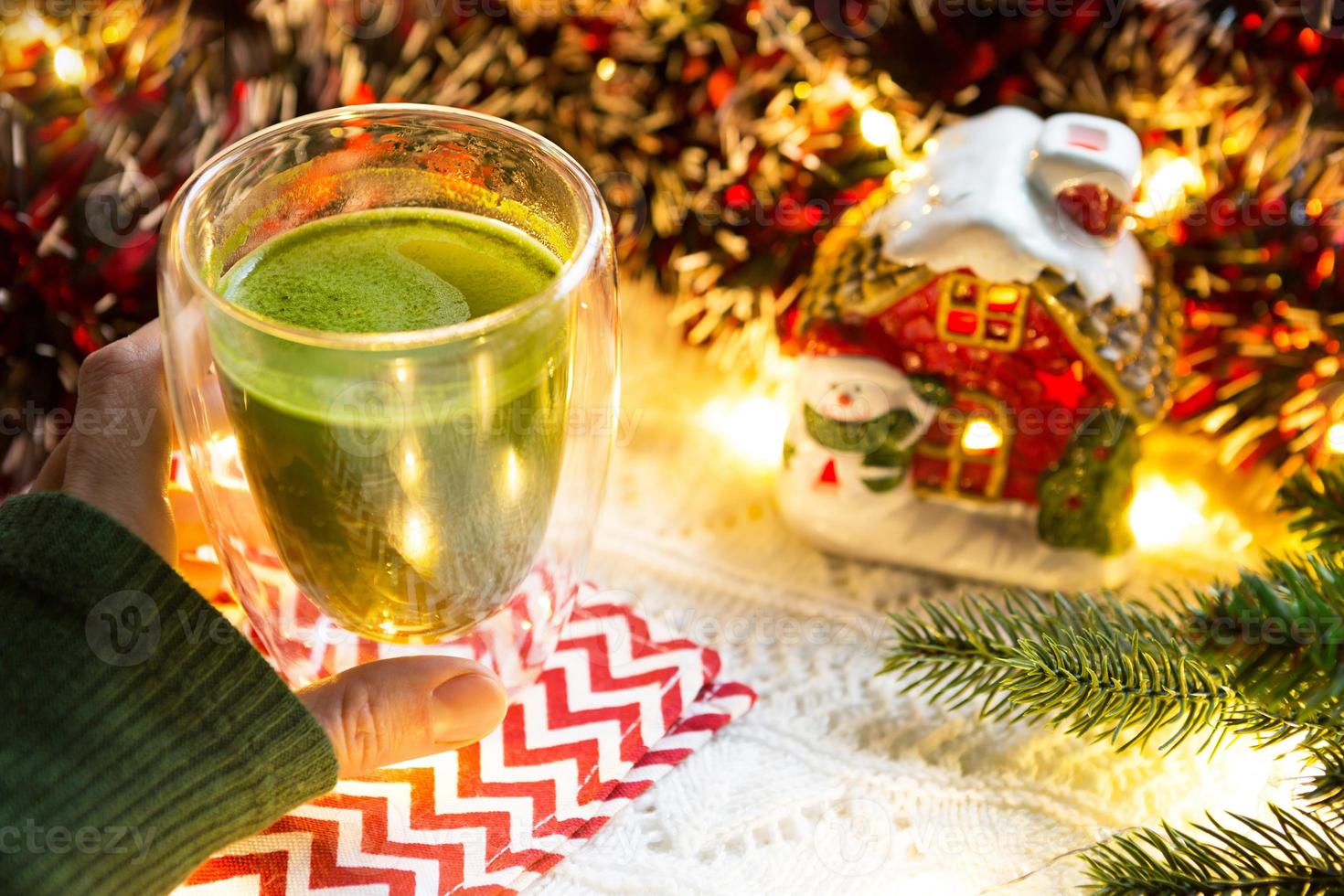 a mão da mulher segura um copo de vidro de parede dupla com chá matcha japonês na mesa com decoração de natal. atmosfera de ano novo, guirlanda e enfeites, ramo de abeto, aconchegante, cobertor de malha, guardanapo listrado foto