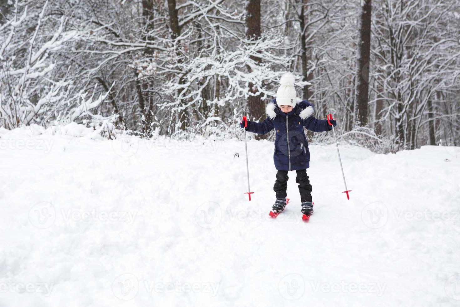 os pés das crianças em esquis de plástico vermelho com bastões atravessam a neve de um escorregador - um esporte de inverno, entretenimento familiar ao ar livre. uma menina desce a encosta desde tenra idade. espaço de cópia foto