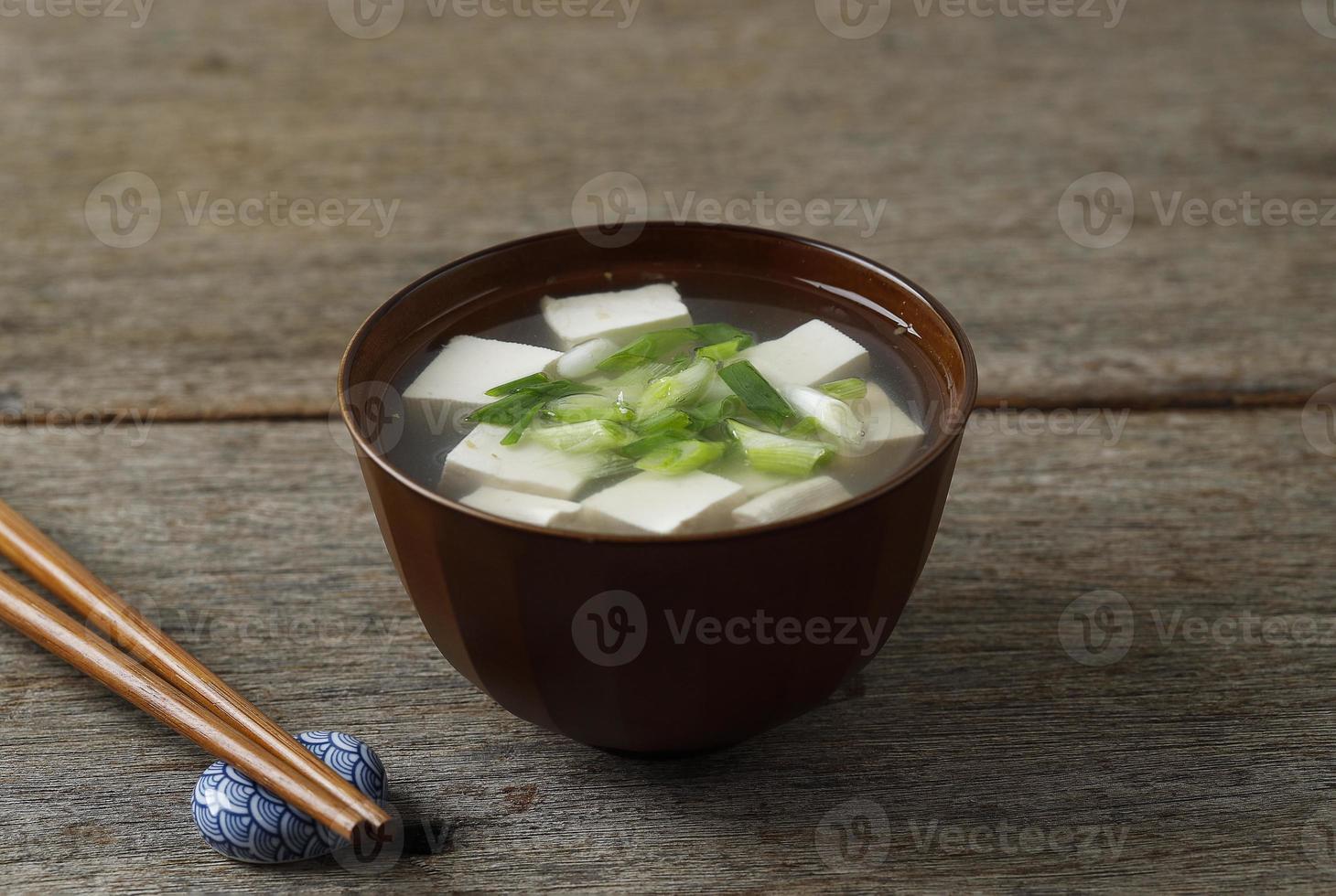 sopa de missô japonesa com cebolinha e queijo tofu em cubos, em uma tigela marrom, mesa de madeira foto