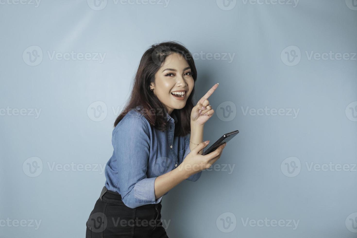 mulher asiática animada vestindo camisa azul apontando para o espaço de cópia ao lado dela enquanto segura um smartphone, isolado por fundo azul foto