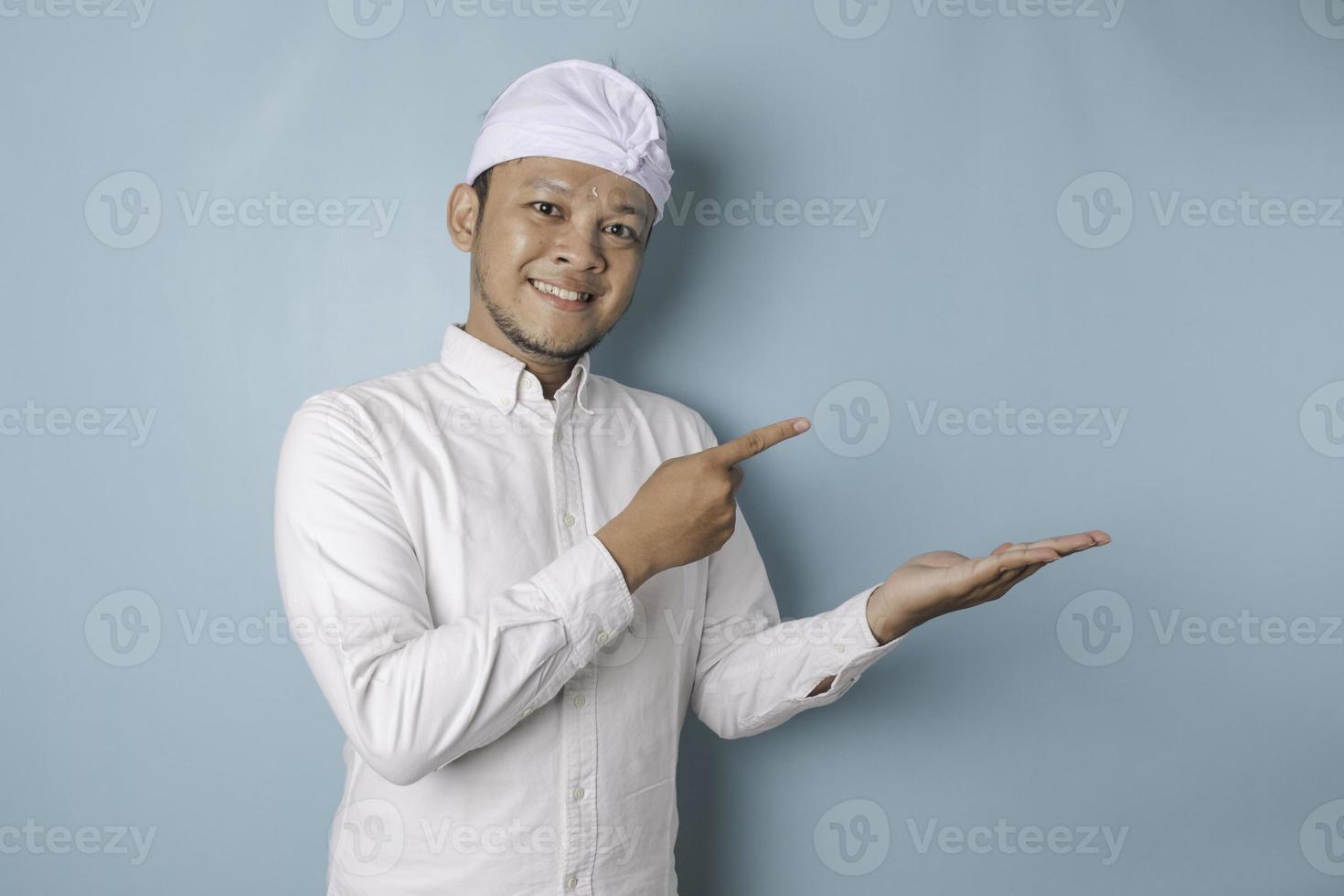 animado homem balinês vestindo udeng ou headband tradicional e camisa branca apontando para o espaço de cópia ao lado dele, isolado por fundo azul foto