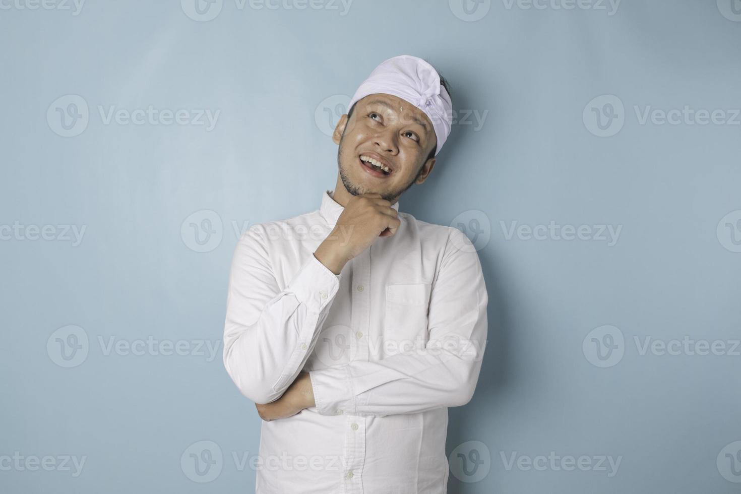 retrato de um jovem balinês pensativo vestindo udeng ou bandana tradicional e camisa branca, olhando de lado enquanto o dedo no queixo isolado sobre fundo azul foto