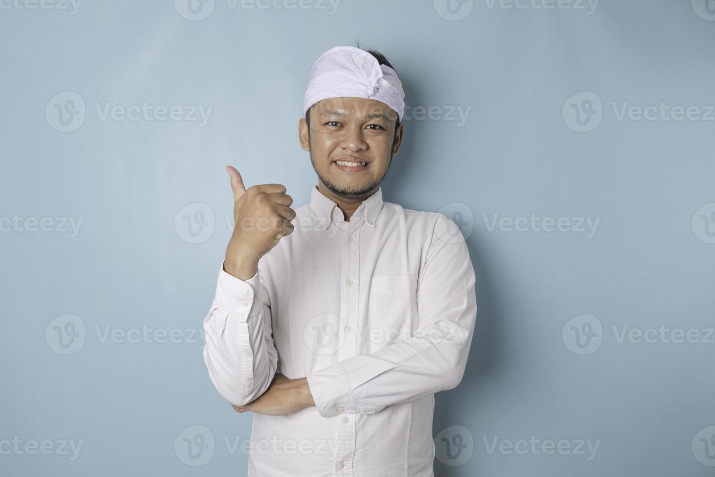 animado homem balinês vestindo udeng ou headband tradicional e camisa branca dá polegares para cima gesto de aprovação, isolado por fundo azul foto