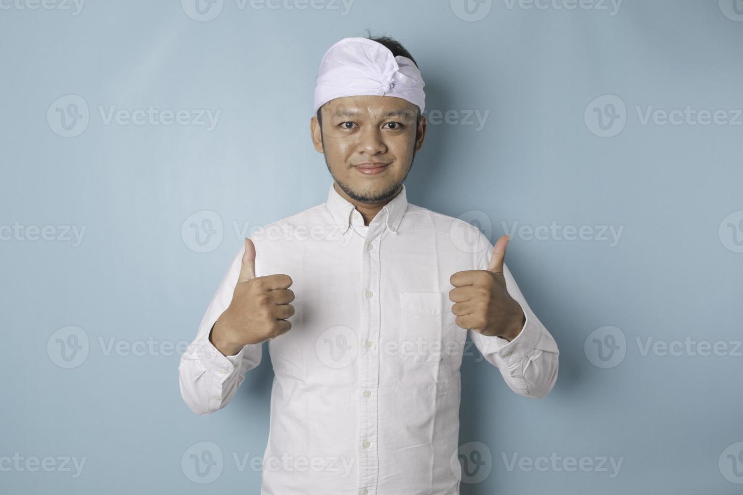 animado homem balinês vestindo udeng ou headband tradicional e camisa branca dá polegares para cima gesto de aprovação, isolado por fundo azul foto