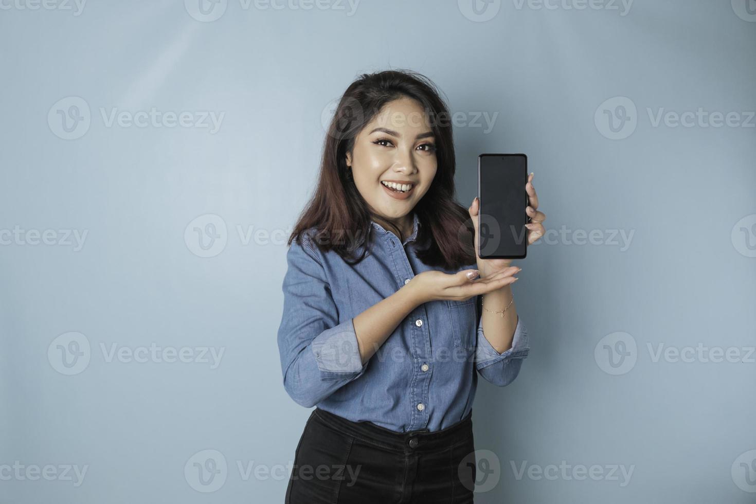 mulher asiática animada vestindo camisa azul apontando para o espaço de cópia em seu smartphone, isolado por fundo azul foto