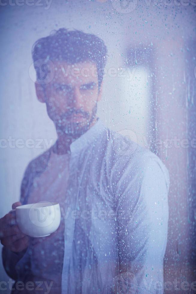 jovem relaxado bebe primeiro café da manhã com gotas de chuva na janela foto