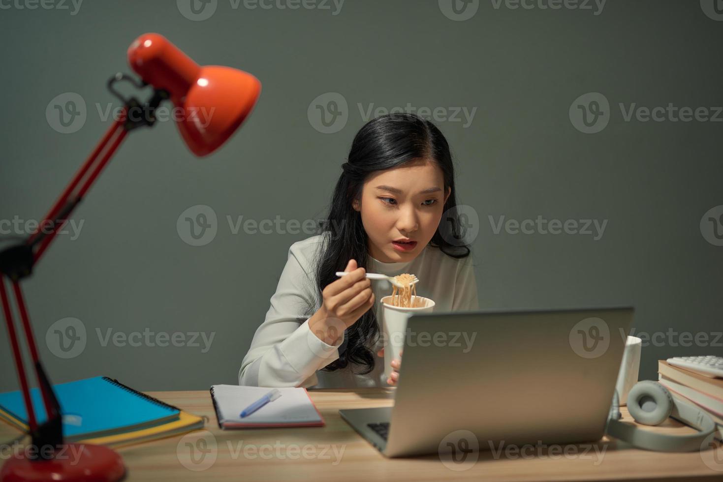 mulheres de negócios inteligentes freelance da ásia comendo macarrão instantâneo enquanto trabalhava no laptop na sala de estar em casa à noite foto