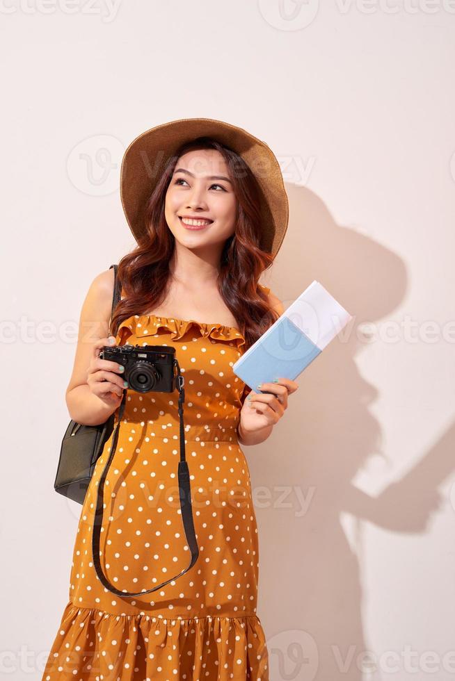 retrato de uma jovem feliz no chapéu segurando a câmera e mostrando o passaporte em pé isolado sobre fundo bege foto
