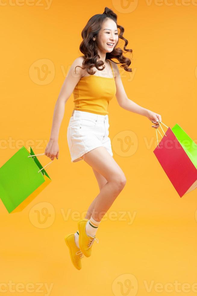 atraente feliz sorridente mulher elegante viciada em compras pulando correndo segurando sacolas de compras em fundo amarelo isolado foto