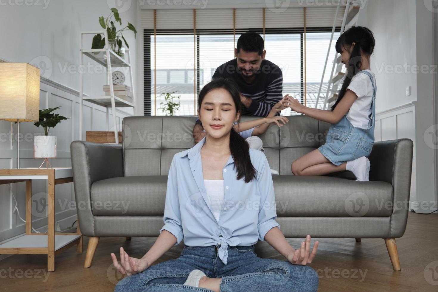 jovem mãe tailandesa asiática senta-se no chão da sala de estar, medita e pratica ioga para saúde e bem-estar quando pai e filhos brincam juntos no sofá, estilo de vida doméstico feliz no fim de semana em família. foto