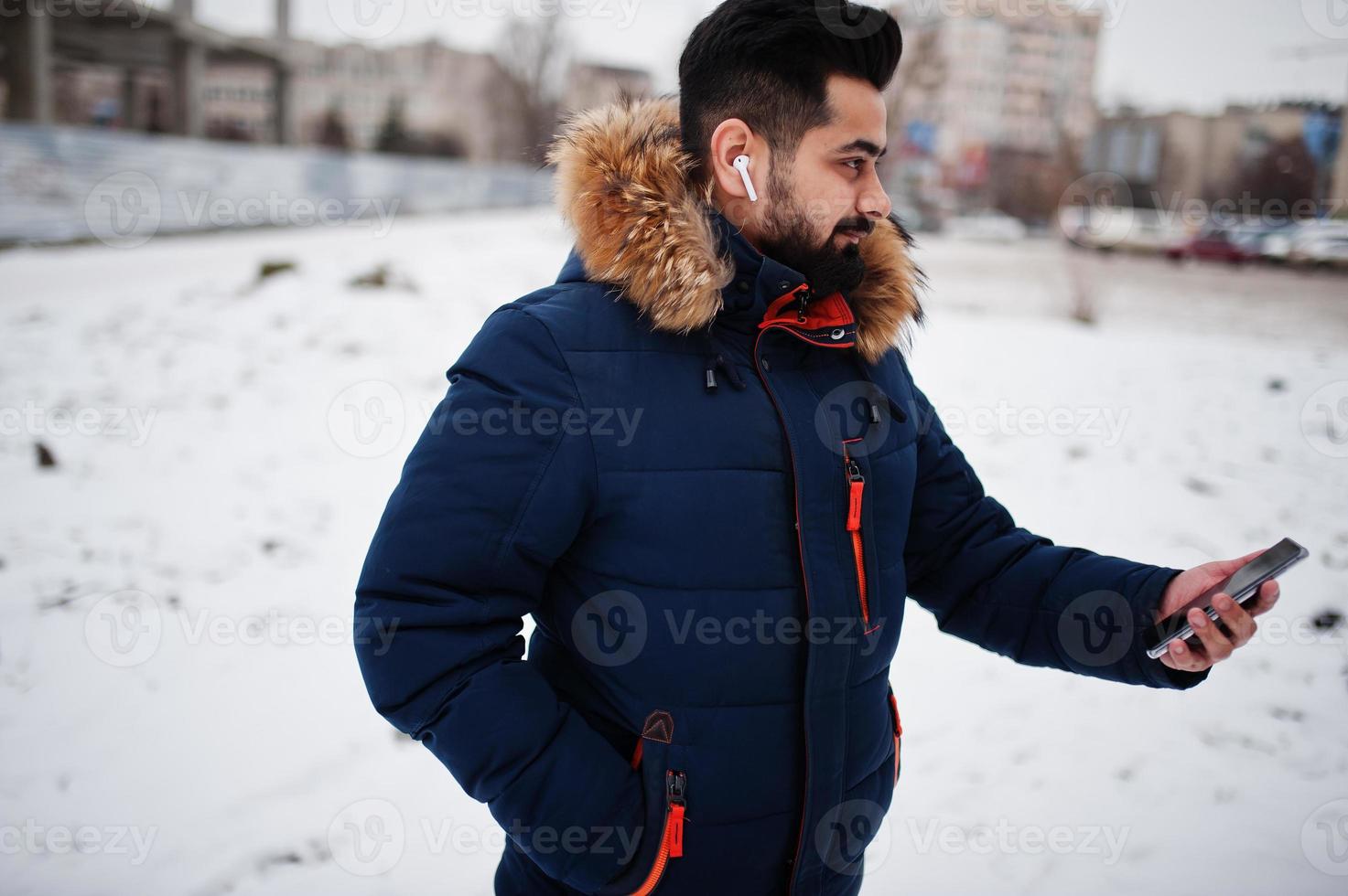 homem indiano de barba usa jaqueta em dia frio de inverno. ele fala celular por fones de ouvido sem fio. foto