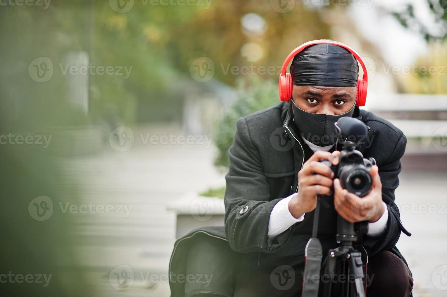 jovem cinegrafista americano africano profissional segurando câmera profissional com equipamento profissional. cinegrafista afro vestindo duraq preto e máscara de proteção facial, fazendo vídeos. foto