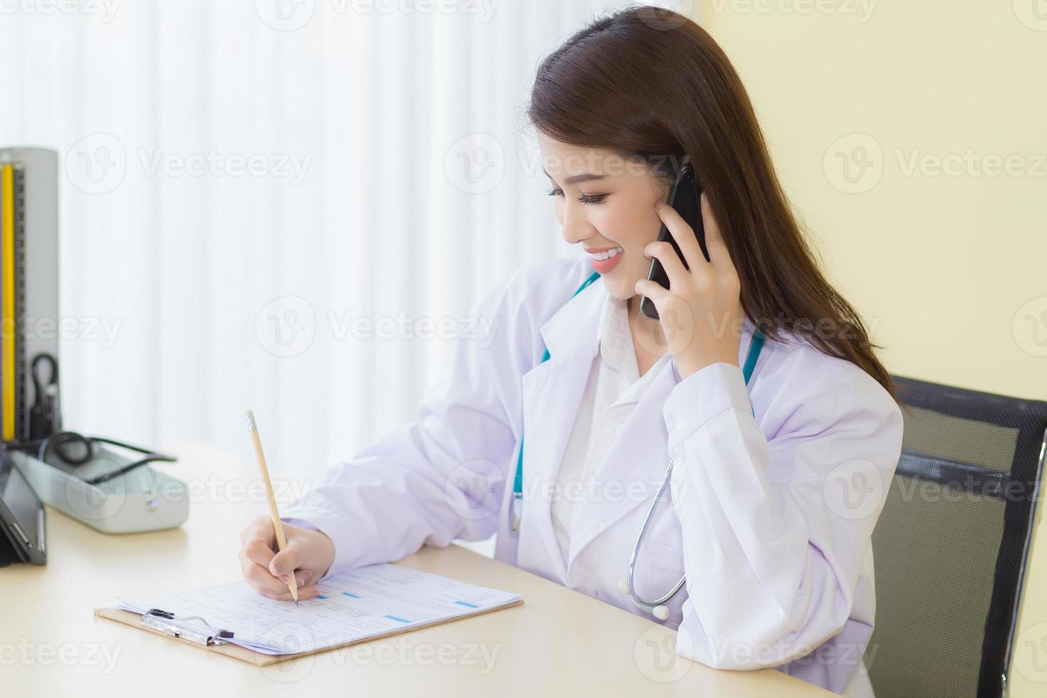 conceito de proteção contra coronavírus. bela médica asiática sentada sorrindo vestindo uma túnica branca e estetoscópio enquanto fornece consulta por telefone foto