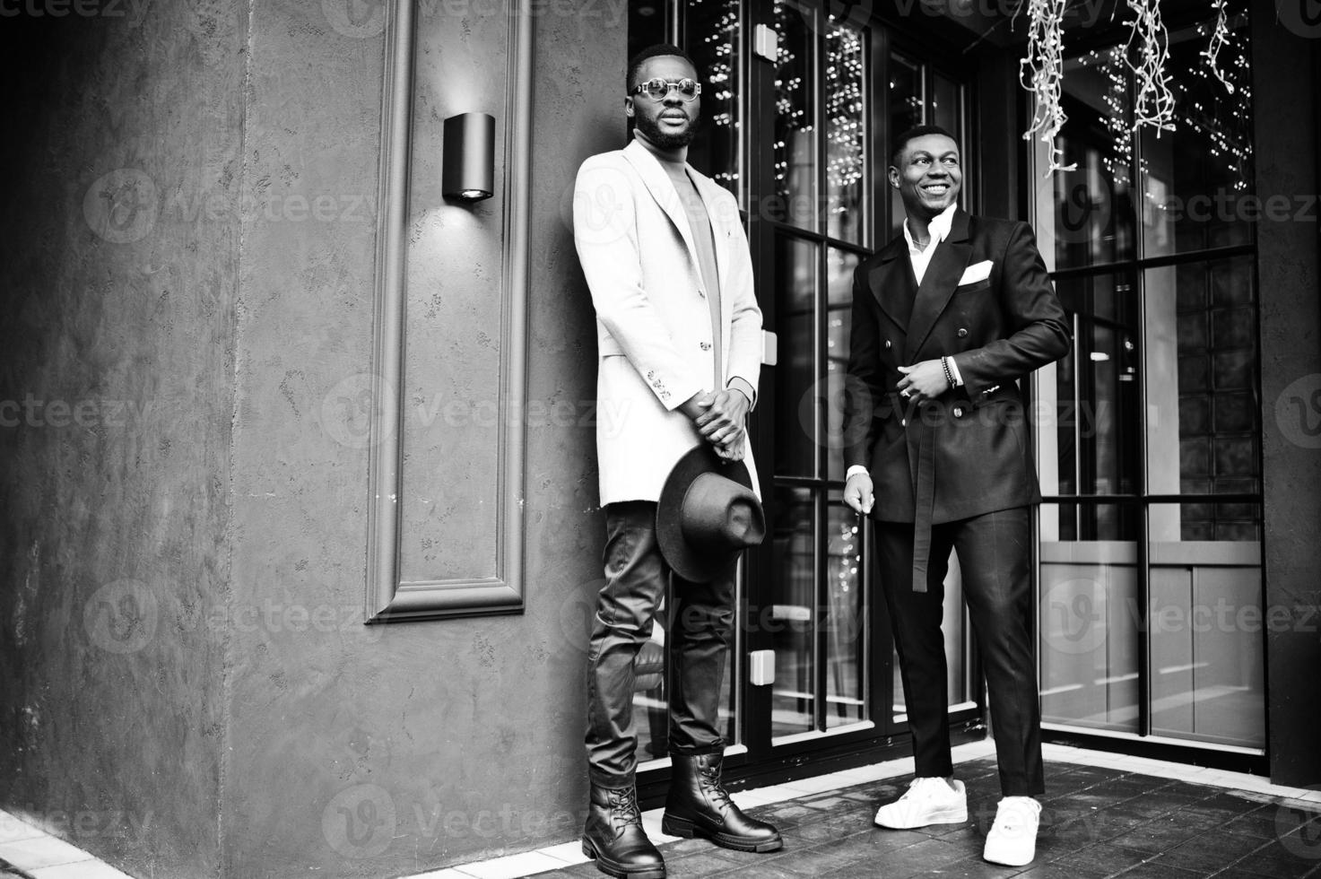dois homens negros da moda posam contra a casa com guirlandas. retrato elegante de modelos masculinos afro-americanos. usar terno, casaco e chapéu. foto