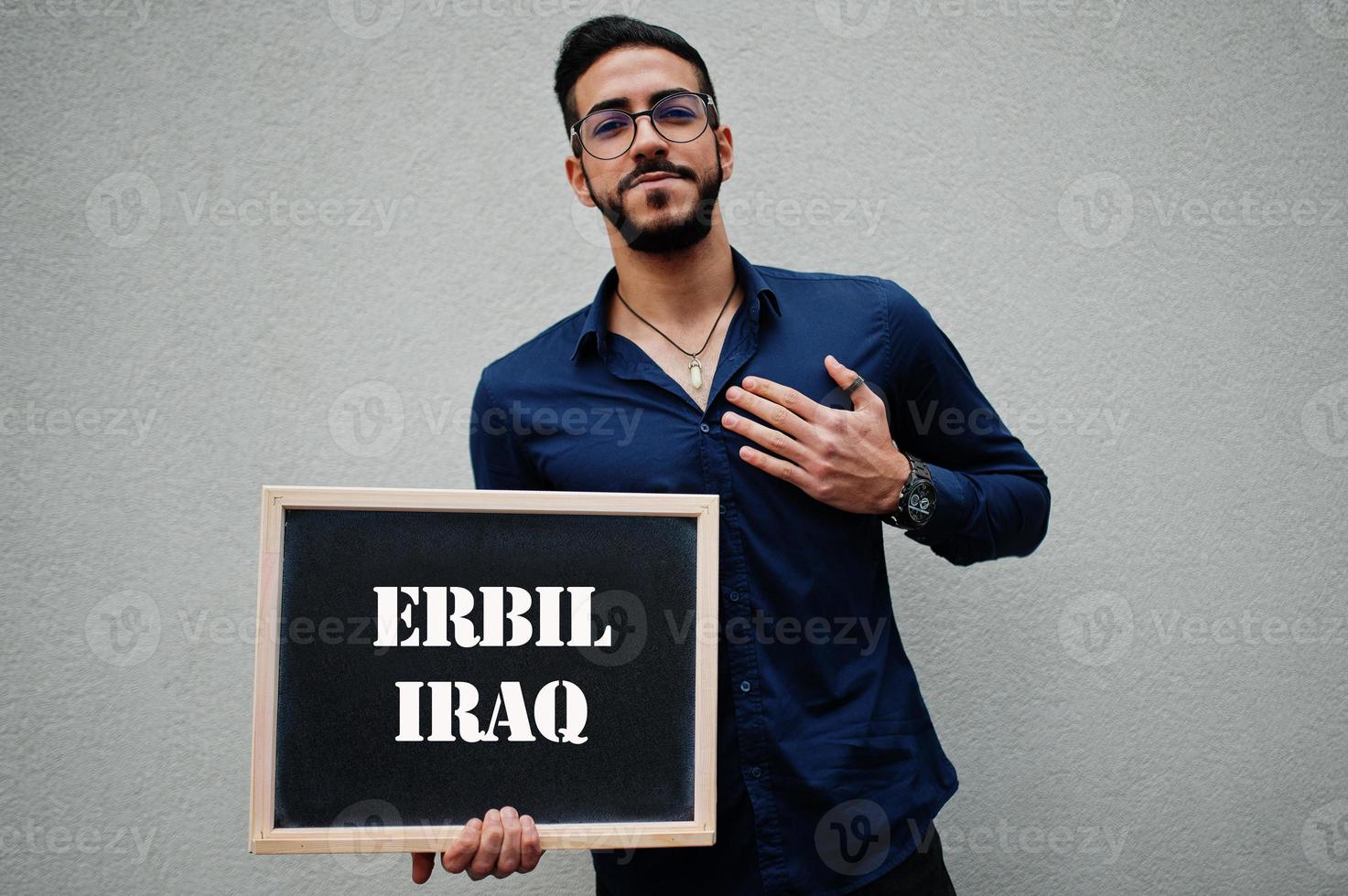 homem árabe usa camisa azul e óculos segura placa com inscrição erbil iraque. maiores cidades no conceito do mundo islâmico. foto