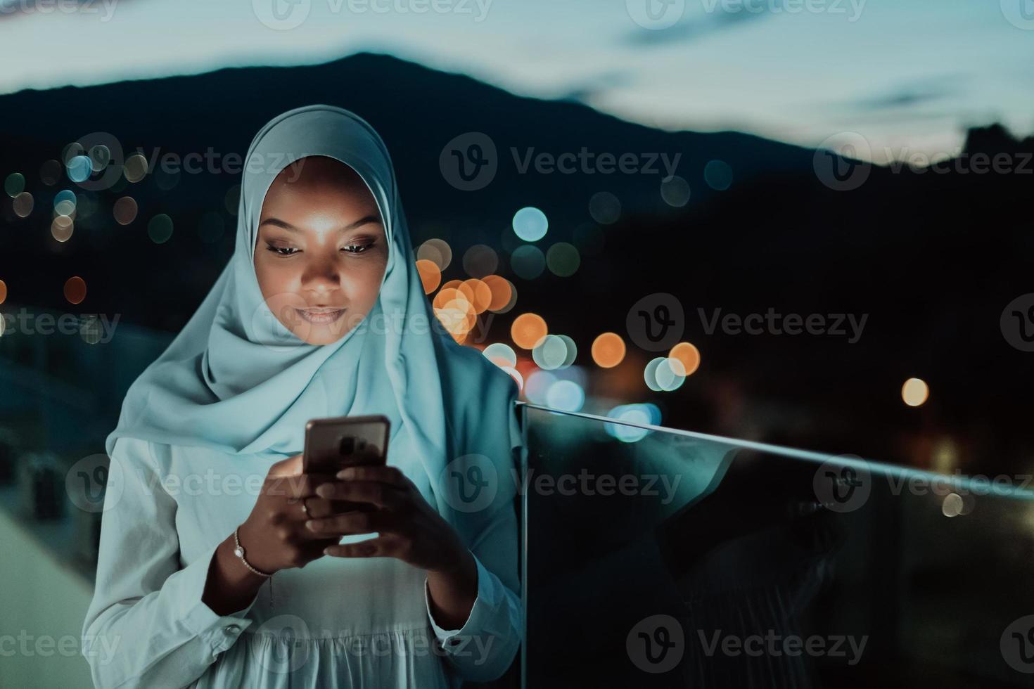 jovem muçulmana vestindo véu de cachecol na rua urbana da cidade à noite enviando mensagens de texto em um smartphone com luz da cidade bokeh ao fundo. foto