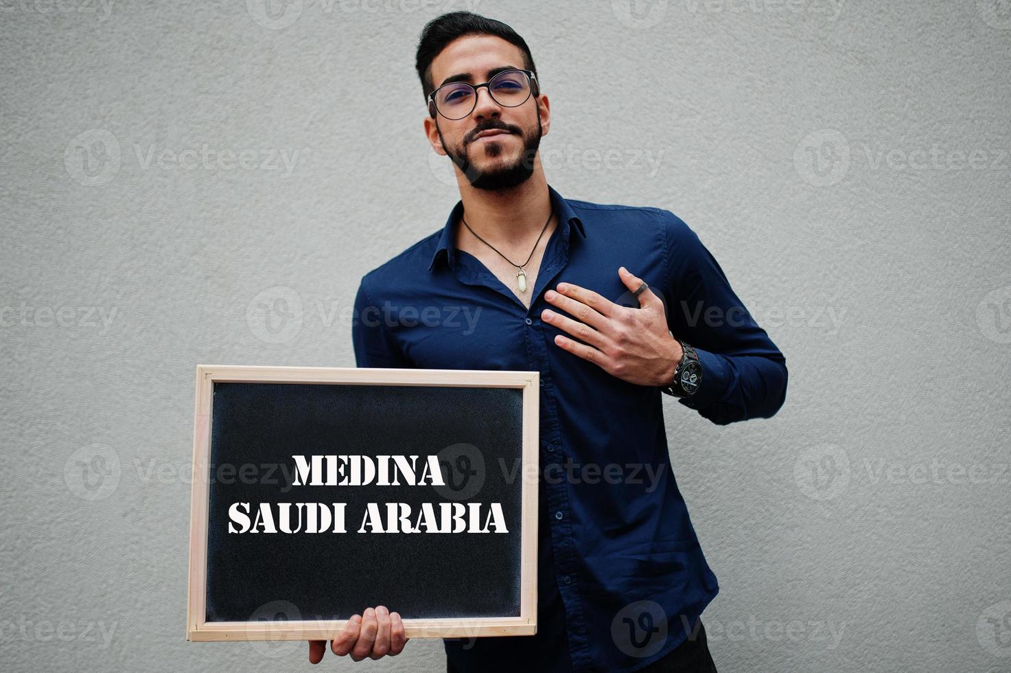 homem árabe usa camisa azul e óculos segura placa com inscrição medina arábia saudita. maiores cidades no conceito do mundo islâmico. foto