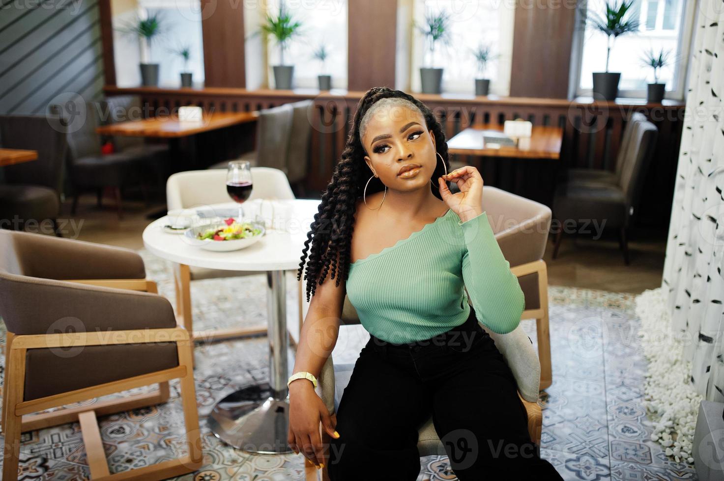 retrato de mulher jovem e atraente afro-americana vestindo suéter verde e calça jeans preta posar no restaurante. foto