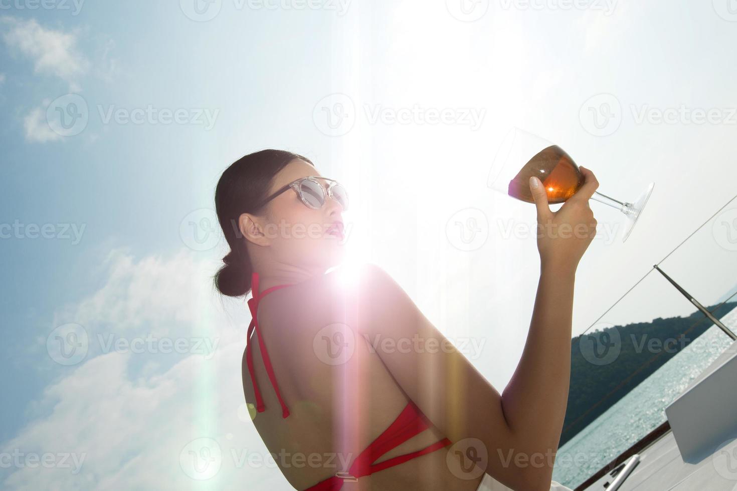 linda mulher de pele bronzeada de raça mista asiática anda ao longo de iates de luxo no oceano profundo, garota de óculos de sol de biquíni vermelho posando como modelo no cais de ancoragem sob o céu azul de verão nas férias foto