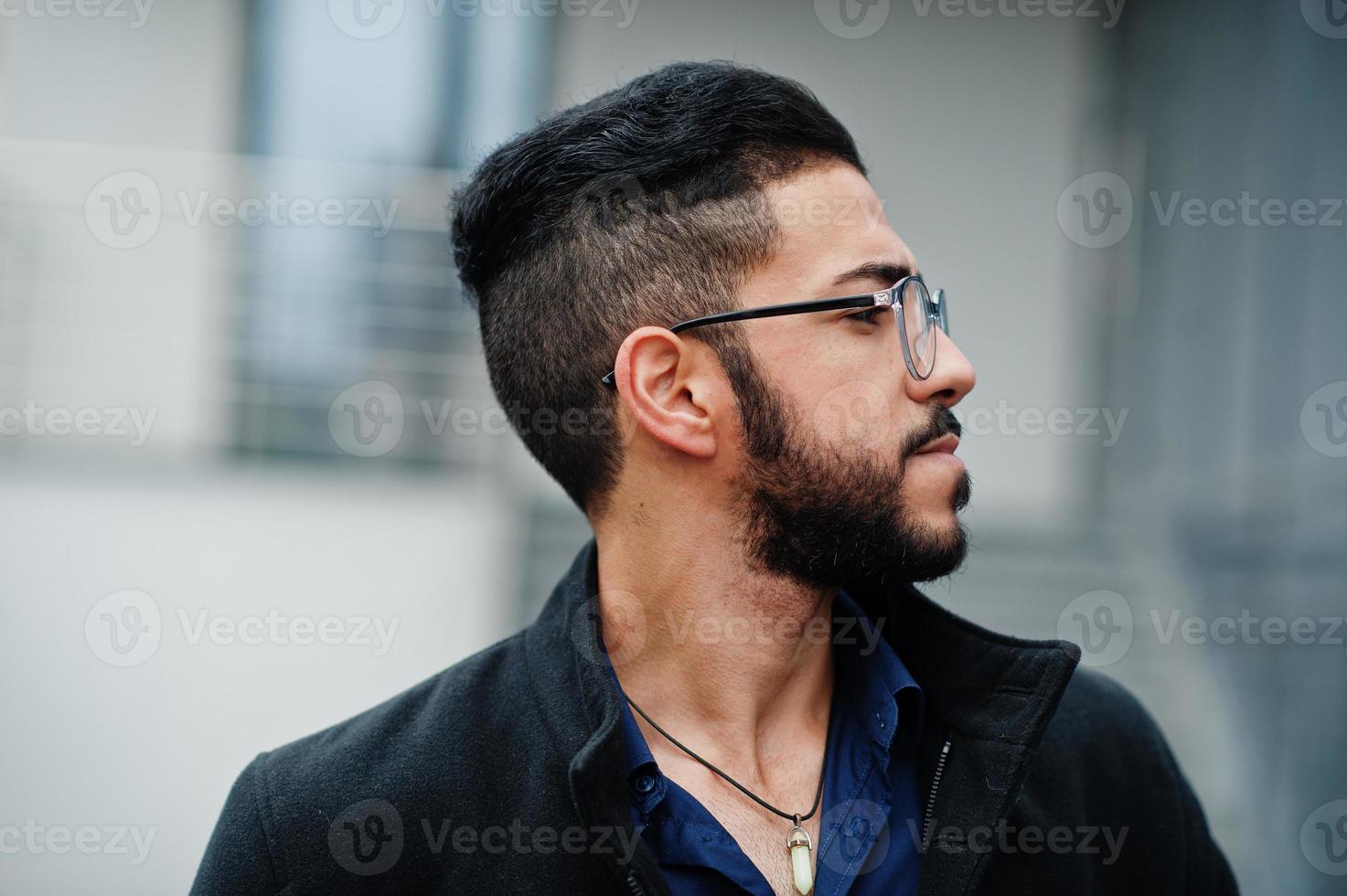 Feche o retrato do empresário de barba do Oriente Médio vestindo casaco preto e camisa azul, óculos contra prédio de escritórios. foto