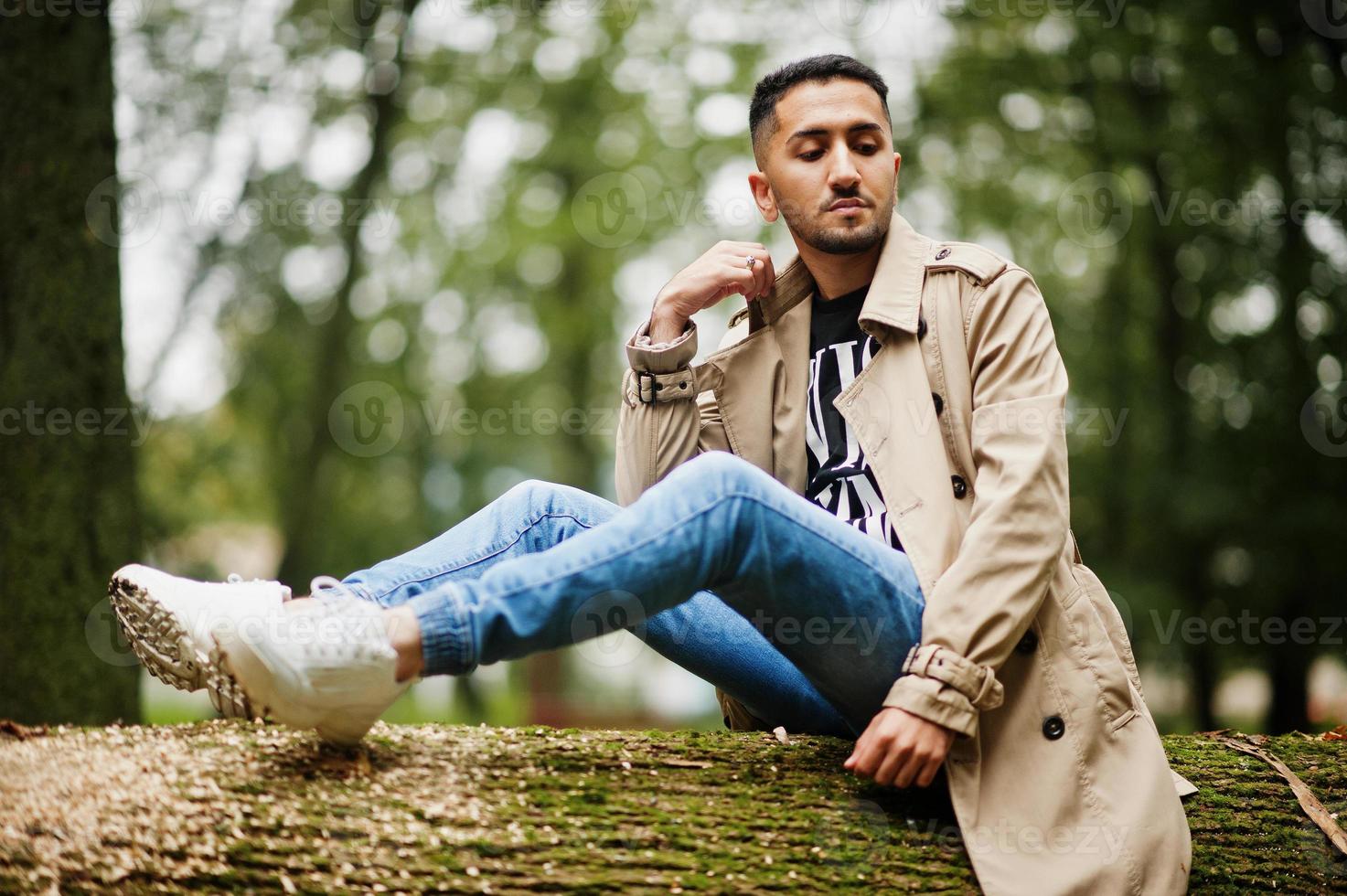 homem elegante kuwaitiano no trench coat sente-se na árvore na madeira. foto