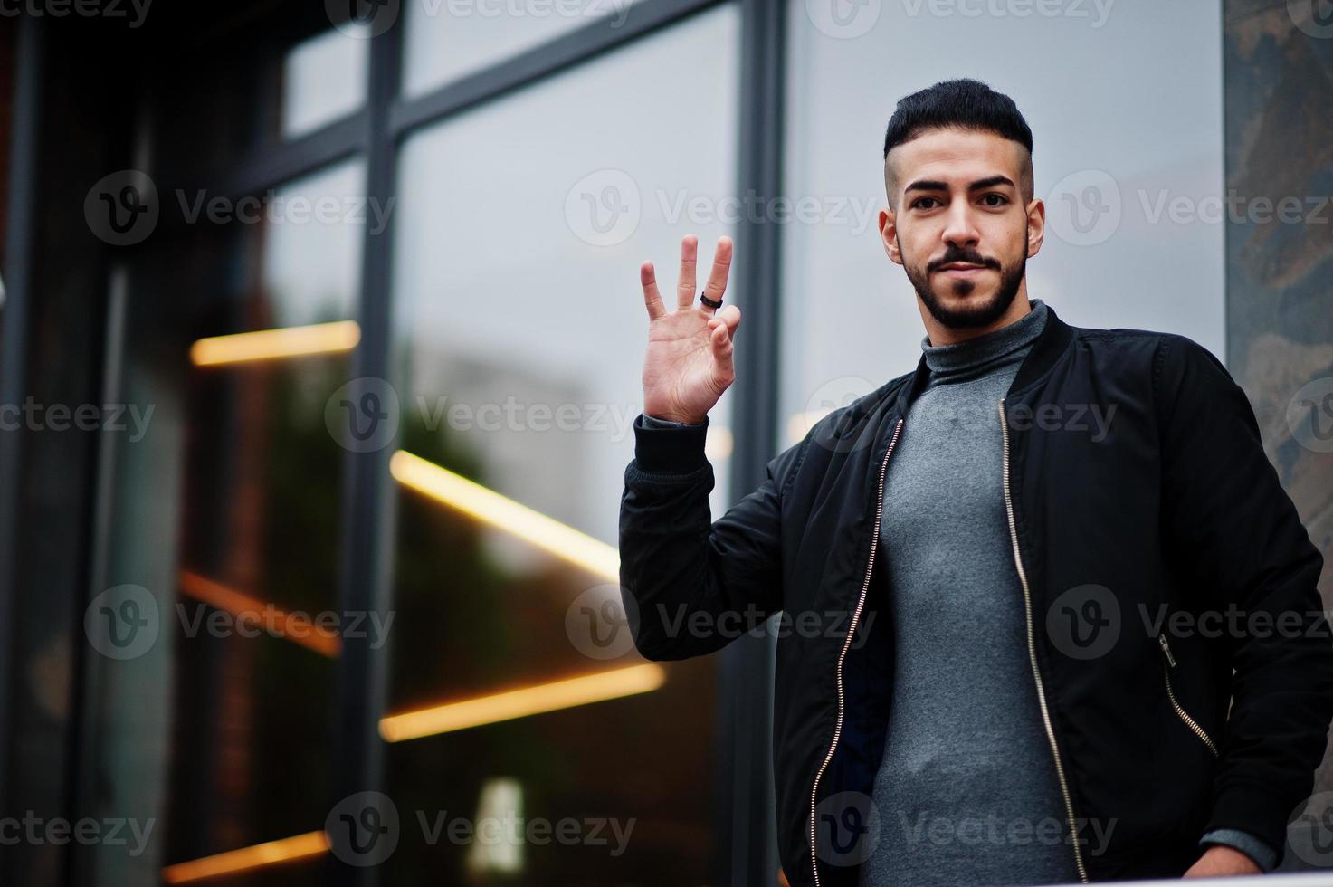 retrato de homem de barba árabe elegante usar gola alta cinza e jaqueta preta. cara modelo árabe mostra três dedos. foto