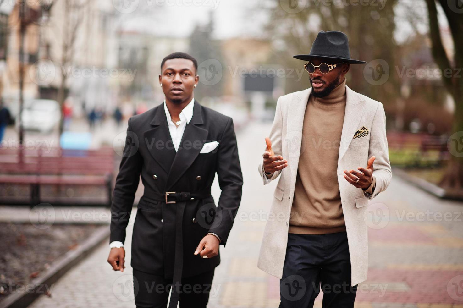dois homens negros da moda andando na rua. retrato elegante de modelos masculinos afro-americanos. usar terno, casaco e chapéu. foto