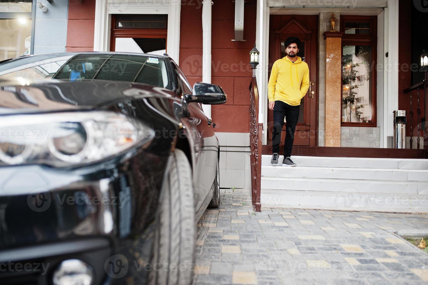 homem indiano urbano jovem hippie em um moletom amarelo na moda. cara legal do sul da ásia usa moletom na varanda da casa com carro de negócios preto. foto