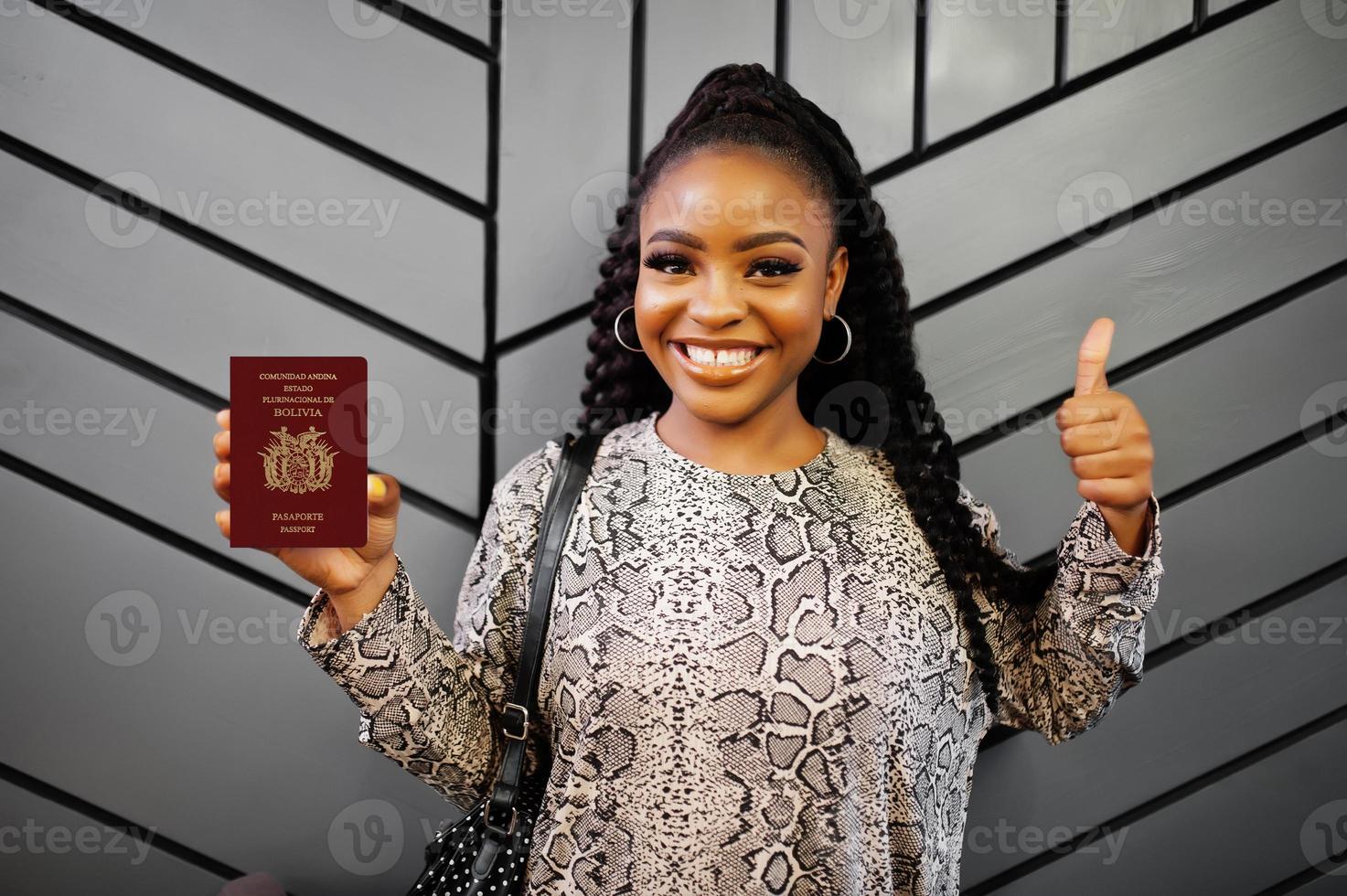 Feche o retrato de uma jovem afro-americana positiva segurando o passaporte da Bolívia e polegares para cima. foto