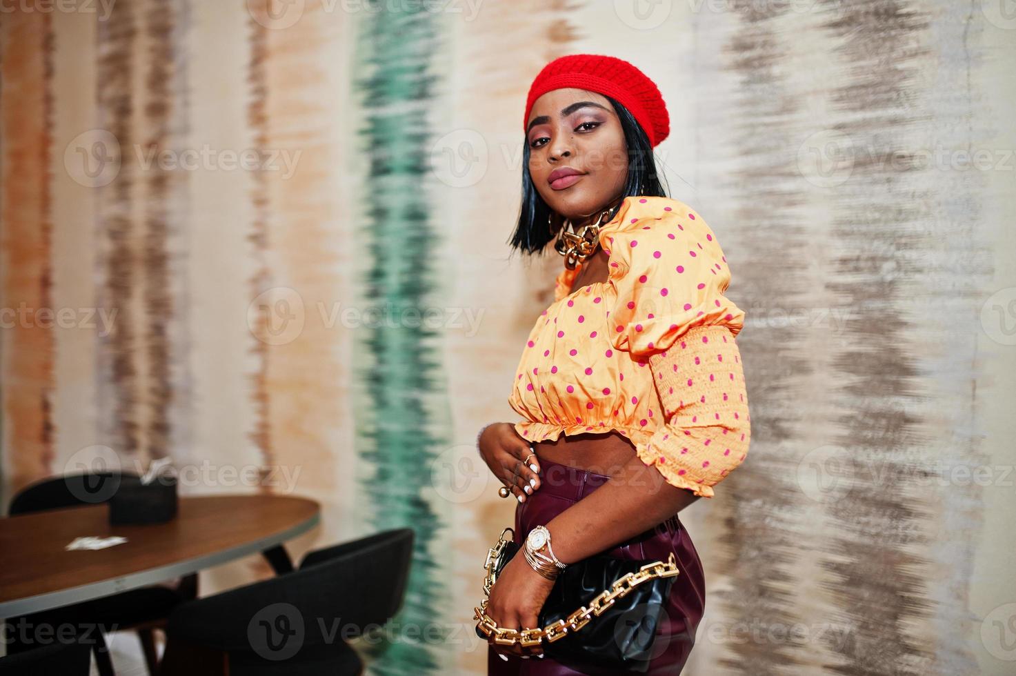 elegante mulher afro-americana de boina vermelha vermelha, blusa de bolinhas de corrente de pescoço de ouro grande e calças de couro posam no interior. foto