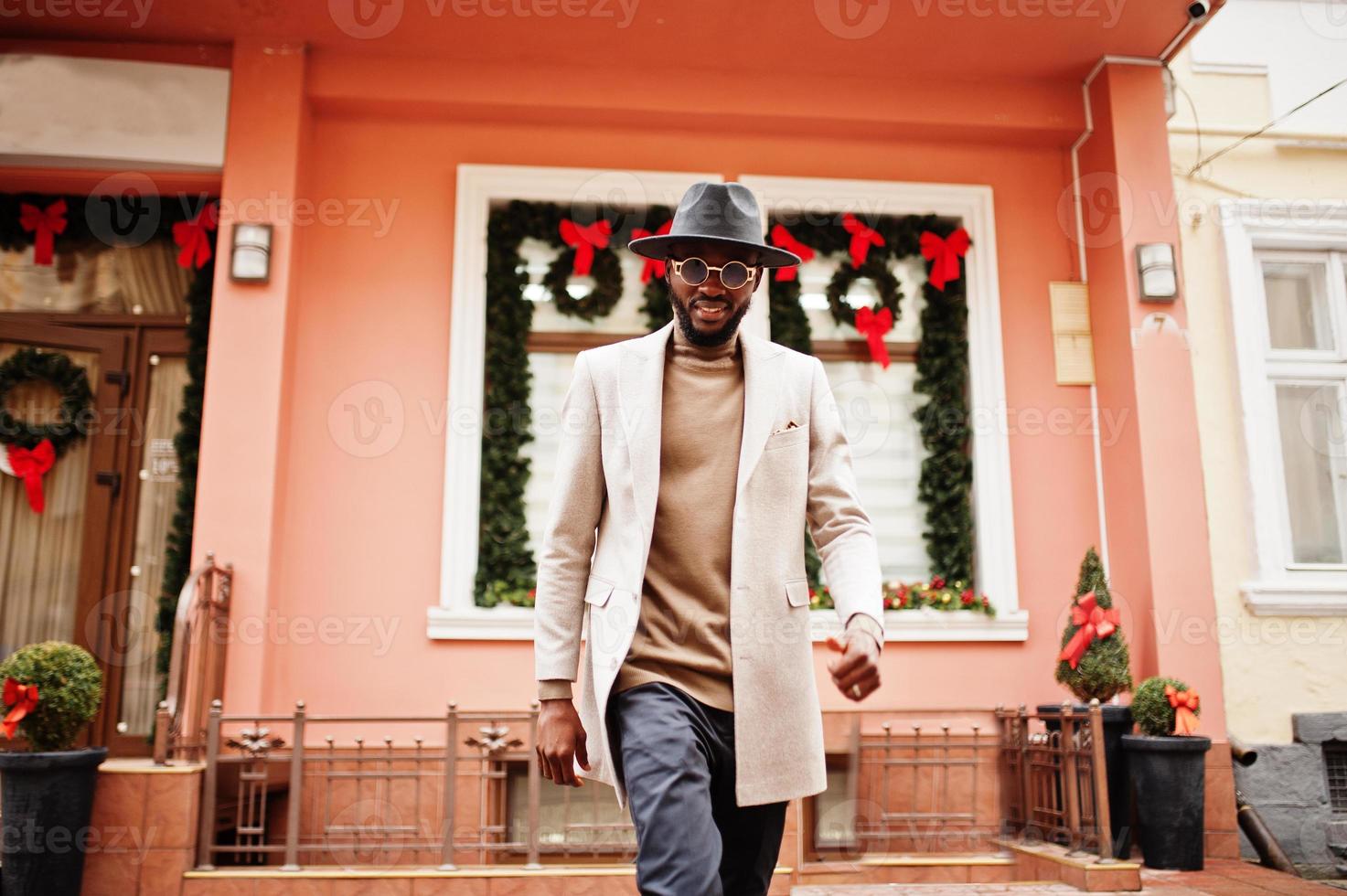 homem afro-americano elegante usa jaqueta bege e chapéu preto com óculos de sol posar contra casa com coroa e decorações de ano novo. foto