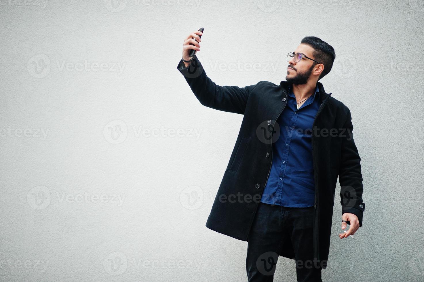 empresário do Oriente Médio usa casaco preto e camisa azul, óculos em pé contra a parede branca, fumando cigarro e fazendo selfie por telefone. foto