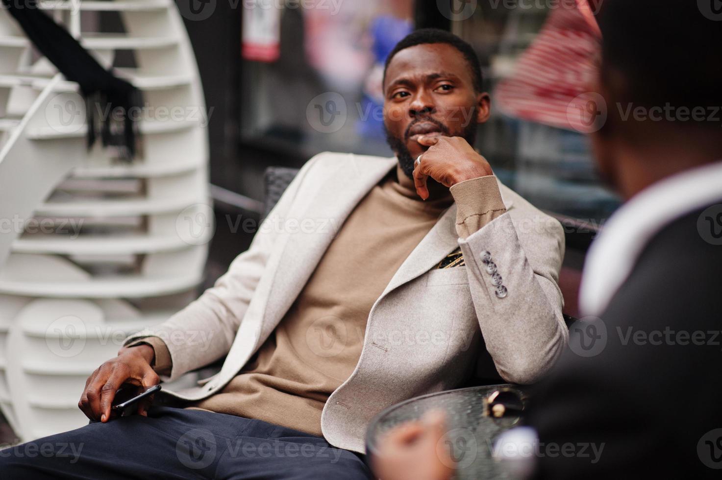 dois homens negros de moda sentados ao ar livre com telefone celular. retrato elegante de modelos masculinos afro-americanos. usar terno, casaco e chapéu. foto